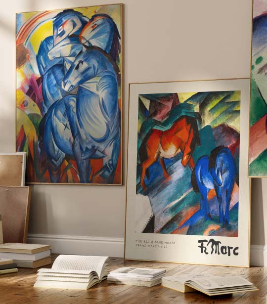 Zwei gerahmte Wandbilder der Bewegung „Der Blaue Reiter“ stehen an einer Wand, davor liegen Bücher auf dem Boden.